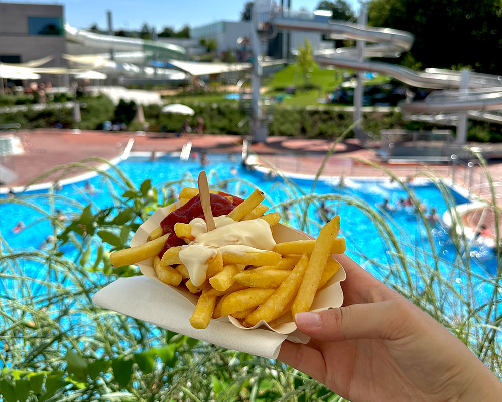 Pommes mit Ketchup und Mayo, im Hintergrund das Freibad Neumarkt