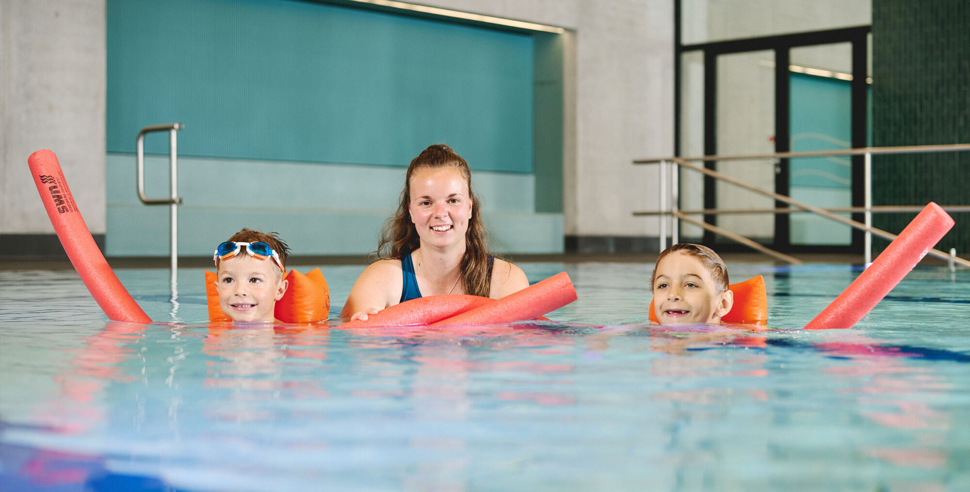 Kursleiterin mit zwei Jungen, die mit Schwimmflügeln und Schwimmnudeln schwimmen lernen